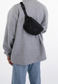 Černý ledvinka se zipovým uzávěrem, připevněný k šedému svetru s nadměrnými rukávy. Hladký povrch, lesklé kovové detaily.