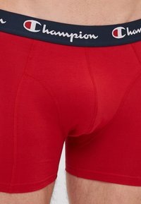 Crvene pamučne bokserice s tamnoplavim pojasom na kojem je bijeli "Champion" logo. Dizajn uključuje šavove za prilagođeni izgled i mekani materijal.