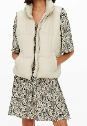 Femme portant un gilet matelassé beige sur une robe courte à manches courtes à motifs, les mains dans les poches du gilet sur fond blanc.