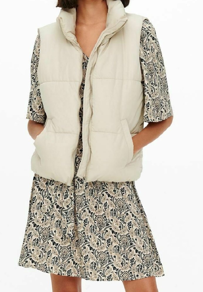 Femme portant un gilet matelassé beige sur une robe courte à manches courtes à motifs, les mains dans les poches du gilet sur fond blanc.
