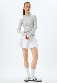 Lys grå langærmet bluse med høj krave, parret med en hvid plisseret nederdel. Modellen bærer hvide sneakers med grå og sorte detaljer.