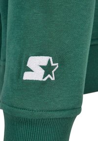 Tessuto felpato verde con una morbida texture, caratterizzato da un logo ricamato bianco con un design di stella e polsini a coste.