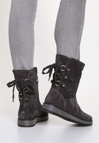 Stivaletti alla caviglia in suede nero con dettagli in pizzo, occhielli dorati e suola texturizzata. Presentano una zip laterale e un tacco piatto e spesso.