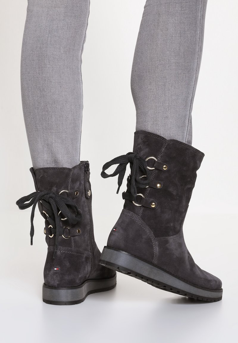 Stivaletti alla caviglia in suede nero con dettagli in pizzo, occhielli dorati e suola texturizzata. Presentano una zip laterale e un tacco piatto e spesso.