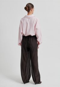 Žena s vlasy svázanými do drdolu, oblečená v volné světle růžové blůze a průsvitných černých kalhotách s širokými nohavicemi, stojící na jednoduchém světle šedém pozadí.