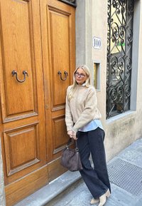 Beige striktrøje lagd over en blå skjorte, parret med sorte brede bukser. Brun håndtaske. Trædør med metalhåndtag og smedejern detaljer.