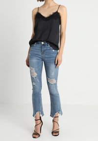 Camisón de satén negro con detalles de encaje, combinado con jeans de denim claro desgastados que tienen dobladillos deshilachados, complementado con tacones negros con tiras.