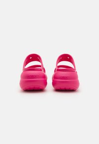 Rožinės Crocs' su kreiva forma ir dviem reguliuojamomis dirželiais; pagamintos iš lengvos medžiagos, turinčios perforacijas oro pralaidumui.