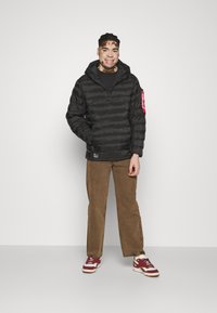 Giacca a vento nera con zip, cuciture orizzontali e un accento rosso sulla manica. Indossata sopra una maglietta nera e abbinata a pantaloni marroni.