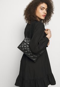 Czarna torba skórzana pleciona z łańcuszkiem, w kształcie prostokąta, o teksturowanej powierzchni, trzymana przez modelkę w czarnej sukience.