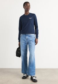 Marine crewneck genser, blå løse jeans, svart handveske og svarte sko med hvite detaljer. Glatt stoff og uformell design.