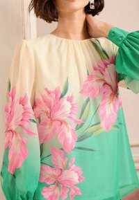 Blusa floreale in tessuto leggero, con grandi fiori rosa su uno sfondo verde sfumato, maniche lunghe a sbuffo e scollatura rotonda.