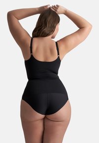 Vrouw met bruin haar die een zwarte tanktop en zwarte hoogglansonderbroek draagt, staand met haar handen achter haar hoofd geheven.