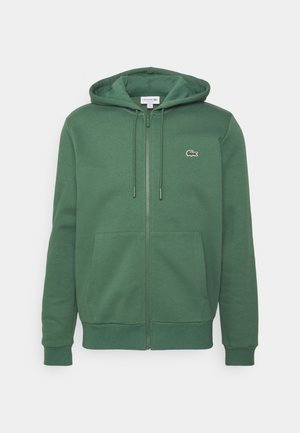 Grüner Hoodie mit Reißverschluss, Fronttaschen, Kapuze mit Kordelzug, gerippten Bündchen und kleinem weißen Lacoste Krokodil-Logo auf der linken Brust.