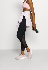 Mustat leggingsit, joissa on verkko-osuuksia, yhdistettynä vaaleanpunaiseen urheiluliivihin ja solmittuun vaaleanpunaiseen paitaan. Jalassa vaaleanpunaiset Nike-tennis kengät ja kädessä musta vesipullo.