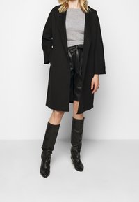 Manteau noir à larges manches et coupe décontractée, porté sur un haut gris et un short en cuir noir, associé à des bottes noires montantes.