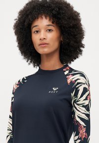 Rash guard à manches longues en bleu marine avec des manches à motif floral, fabriqué à partir d'un matériau extensible. Doté d'un petit logo sur la poitrine.
