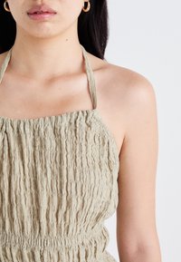 Artimiausias plano vaizdas moters viršutinės kūno dalies, which is wearing a textured, light green halter top with gathered detailing, showcasing her face and earrings.