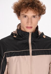 Gevoerde jas met een zwart bovenstuk en een beige onderstuk, gemaakt van lichtgewicht materiaal, met een rits en trekkoorden.