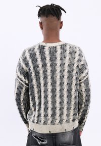 Personne avec des dreadlocks courtes portant des boucles d'oreilles anneaux et un pull en maille à motifs noir et blanc, associé à un jean noir déchiré.