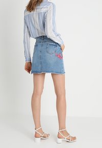 Jupe en denim mini avec ourlet effiloché, design ajusté et broderie florale rose sur la poche arrière. Associée à une chemise rayée bleue et des sandales blanches.