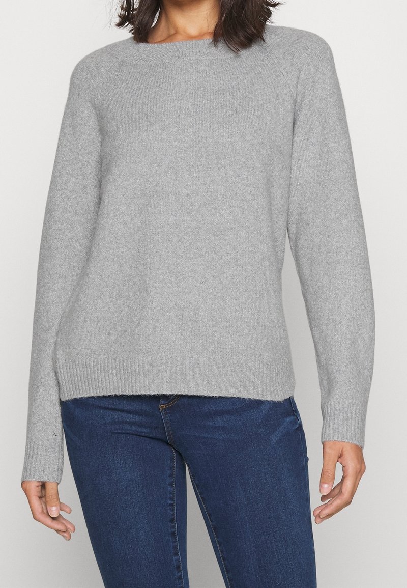 Femme portant un pull léger gris uni à manches longues et un jean bleu foncé, debout devant un fond neutre.