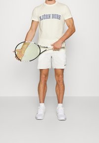 Vit t-shirt med texten "BJÖRN BORG", vita shorts och vita sneakers. Håller en tennisracket med gröna och svarta detaljer.