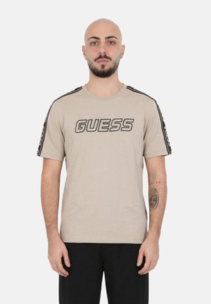 Homme chauve avec barbe portant un t-shirt beige de la marque Guess avec un logo noir et des rayures sur les manches, debout devant un fond blanc uni.