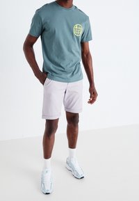 T-shirt en coton teal avec un logo circulaire jaune, shorts gris clair et baskets blanches. Le modèle se tient les mains dans les poches sur un fond uni.