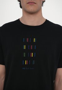 Musta T-paita, jossa on monivärisiä pystysuoria viivoja edessä ja "PS Paul Smith" -logo sinisenä alareunassa. Lyhyet hihat, pyöreä kaula-aukko.