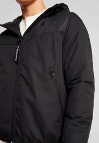 Black padded jacket with a hood and zip closure. Features a chest logo, side pockets, and a smooth matte texture. Full-length sleeves.