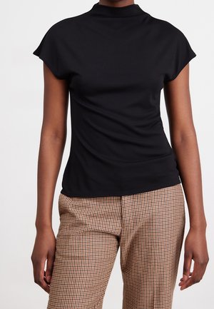 Persona que lleva una camiseta negra ajustada de manga corta con cuello alto y pantalones marrones a cuadros sobre un fondo claro y liso.