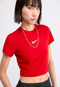 Czerwony, krótki top na ramiączkach wykonany z bawełny, z subtelnym logo Nike w kontrastowym kolorze. Modelka ma na sobie srebrny łańcuszek.