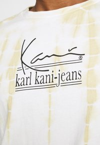 White T-shirt with yellow tie-dye pattern, featuring black printed logo: "Karl Kani Jeans" in cursive and serif fonts.