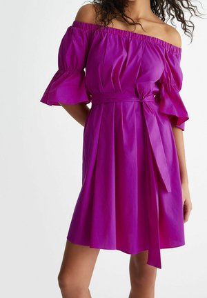 Donna che indossa un vestito viola fino al ginocchio, con le spalle scoperte, maniche arruffate e una cintura annodata in vita, in piedi davanti a uno sfondo semplice.
