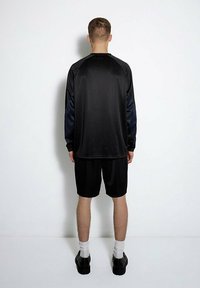 Schwarzes Sportoutfit bestehend aus einem Langarmshirt mit strukturiertem Stoff und Shorts, beide mit lässiger Passform. Ergänzt durch schwarze Schuhe und weiße Socken.