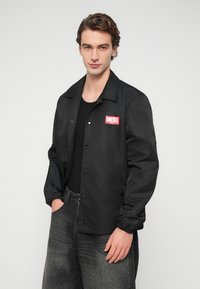 Μαύρο καθημερινό τζάκετ με κουμπιά τύπου snap, ελαστικά τελειώματα στα μανίκια και κόκκινο κεντημένο λογότυπο "DIESEL" στο αριστερό στήθος. Συνδυάζεται με σκουρόχρωμο τζιν παντελόνι.