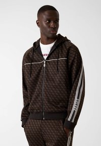 Brązowa bluza z kapturem na suwak z wzorem, białymi paskami i logo "GUESS" na rękawach, wyposażona w kaptur ze sznurkiem oraz ściągacze na mankietach i dole.