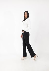 Cardigan blanc à côtes avec col noir, associé à un jean évasé noir et des sandales à talons blanches, sur fond uni.
