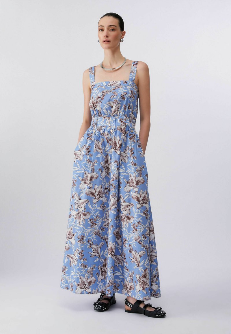 Femme debout, les mains dans les poches, portant une robe maxi sans manches bleu à fleurs et des sandales noires cloutées, sur un fond uni.