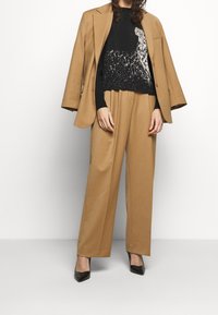 Beżowa marynarka na czarnym swetrze z jasnym wzorem, zestawiona z luźnymi beżowymi spodniami i czarnymi butami z zaostrzonym noskiem. Gładkie faktury w całym stroju.