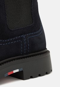 Námornické semišové boty s texturovanou síťovinou, kontrastním modrým šitím, gumovou podrážkou a malým trojbarevným akcentem v blízkosti spodní části.