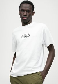 Biela bavlnená tričko s krátkymi rukávmi, na ktorom je umiestnené čierne oválne logo v strede hrude. Model má na sebe olivové nohavice.