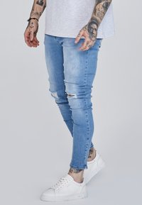 Világoskék skinny farmernadrág, térdnél megviselt részletekkel, fehér sportcipővel párosítva. Az anyag sima textúrájú és testhezálló kialakítású.