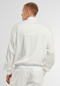 Giacca atletica bianca con una texture liscia, vestibilità rilassata, polsini a costine e colletto, vista da dietro, abbinata a pantaloni coordinati.