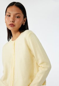 Ljusgul cardigan i mjukt, strukturerat tyg med knäppning fram, rund halsringning och långa ärmar. Minimalistisk design utan mönster.