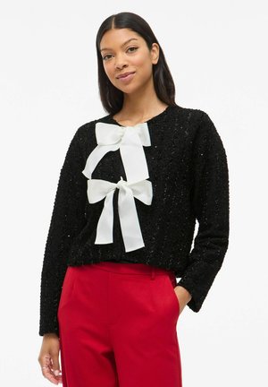 Schwarzer strukturierter Pullover mit einem weißen Bändchen-Detail an der Vorderseite. Der Pullover hat einen runden Halsausschnitt und lange Ärmel. Kombiniert mit roten Hosen.