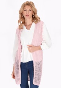 Lichtroze gebreide lange vest met semi-transparante details, gecombineerd met een witte blouse en blauwe jeans. Heeft een open voorkant en geen sluitingen.