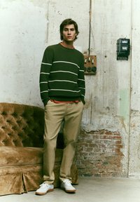 Suéter verde oscuro con rayas blancas, pantalones color beige y zapatillas blancas. El modelo está de pie frente a una pared desgastada, con un sofá de estilo vintage cerca.