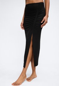 Frau trägt einen schwarzen gerafften Midirock mit einem vorderen Schlitz, steht barfuß vor einem schlichten hellen Hintergrund.